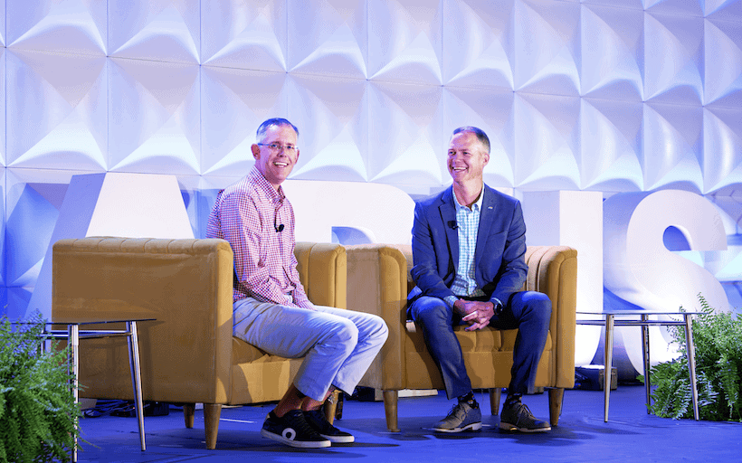 Two men sit and smile on stage in armchairs, engaged in conversation. They are in front of large white letters and geometric wall panels, with ferns and side tables nearby. Both wear microphones.