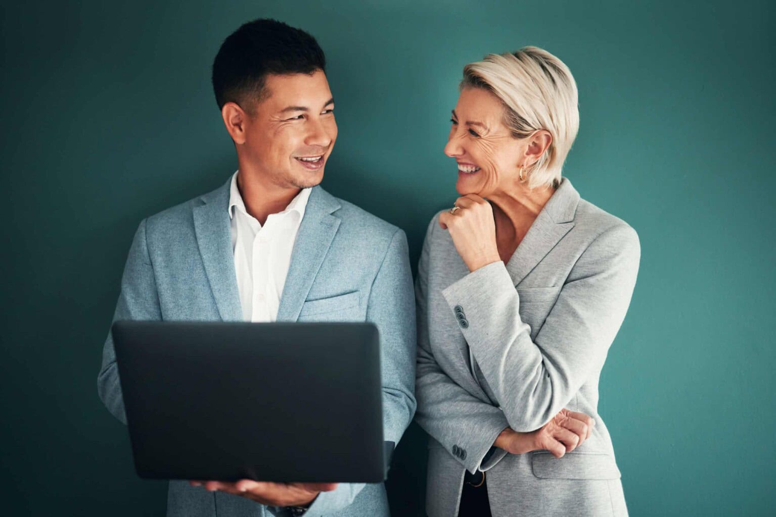 Two professionals in light gray blazers stand against a green background, smiling at each other. One holds an open laptop while the other stands with her hand on her chin, engaged in conversation.