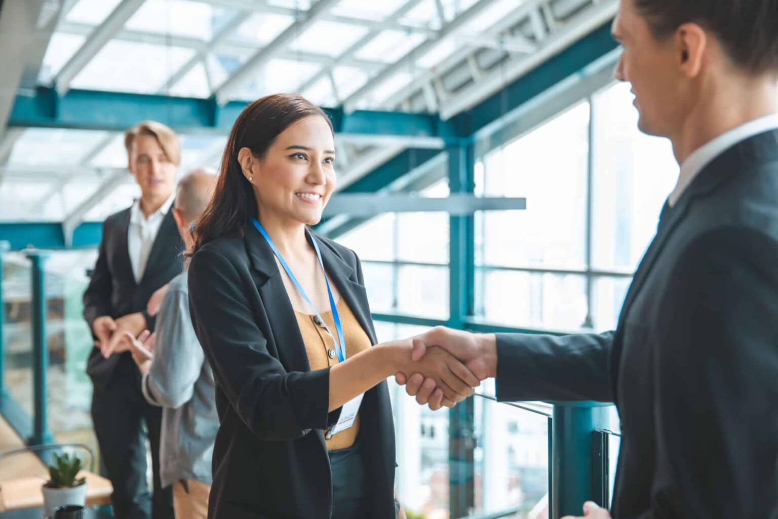 A woman in business attire smiles while shaking hands with a man in an office setting. Two other people stand in the background, and the space is bright with large windows.