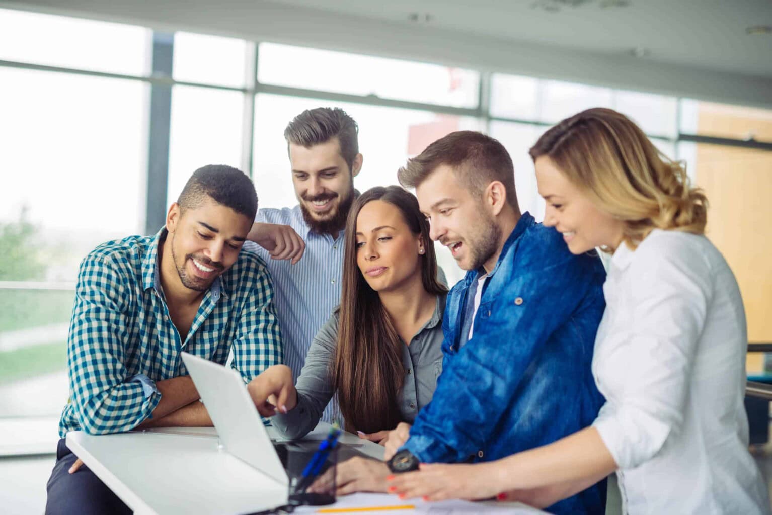Five people gather around a laptop, smiling and looking at the screen, appearing to collaborate on a project in a bright, modern office space with large windows in the background.