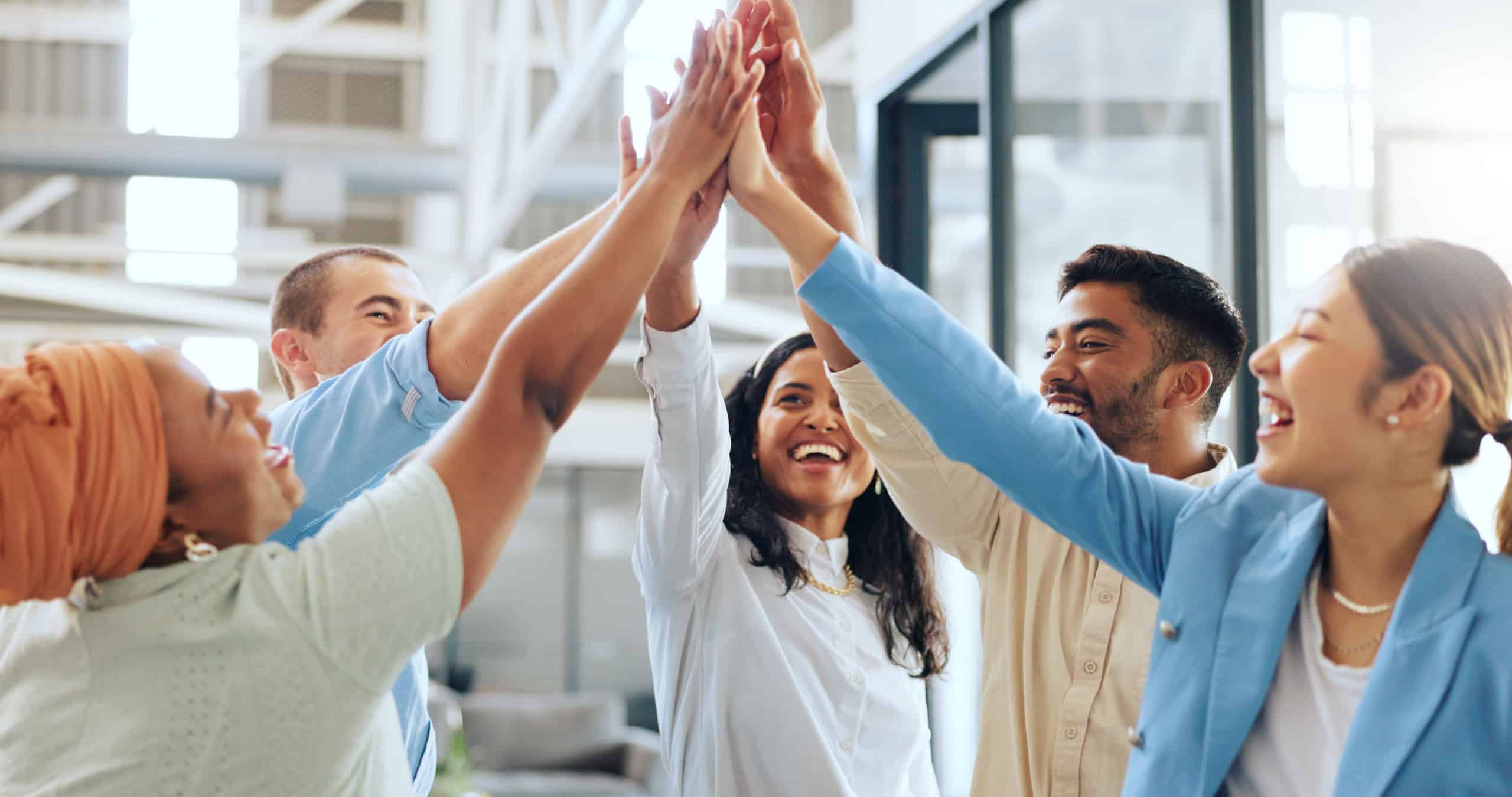 A group of five diverse colleagues stand in a bright office, smiling and giving each other a high-five, celebrating success and teamwork.
