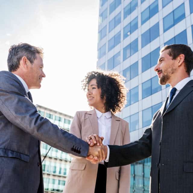 Three businesspeople in formal attire stand outside modern office buildings. Two men are shaking hands while a woman smiles, watching them. The mood appears positive and professional.