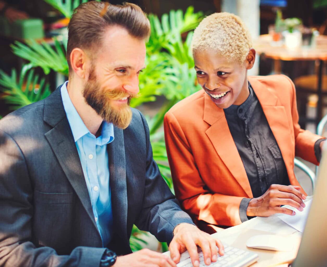 Two professionals, a man and a woman, sit at a desk outdoors, smiling and working together on a computer. Lush green plants surround them, creating a bright and collaborative atmosphere.