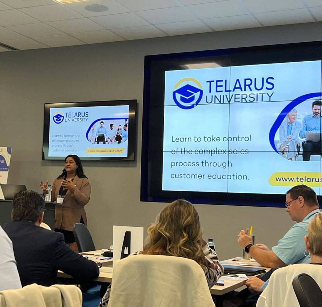 A woman presents at a conference in front of a screen displaying "Telarus University" and the text, "Learn to take control of the complex sales process through customer education." Attendees are seated and listening.