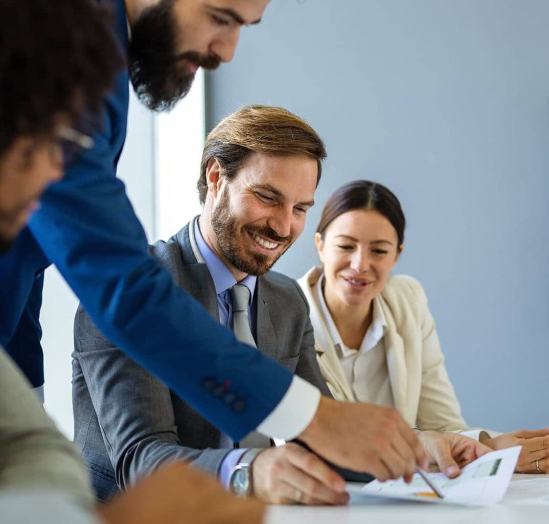 Four professionals in business attire sit at a table, smiling and discussing documents together in a brightly lit office setting. One person is pointing at the papers, leading the conversation.