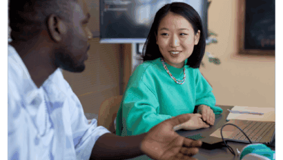 Two people sit at a table, engaged in conversation about data security. A laptop, smartphone, and notebook are on the table with a screen displaying code in the background. The woman is smiling, wearing a green sweater.