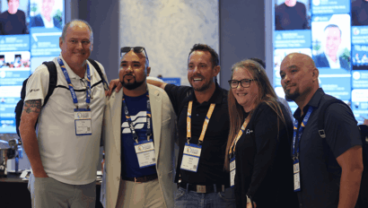 Five people wearing conference badges pose together and smile in a conference setting, with digital screens displaying headshots and information in the background.