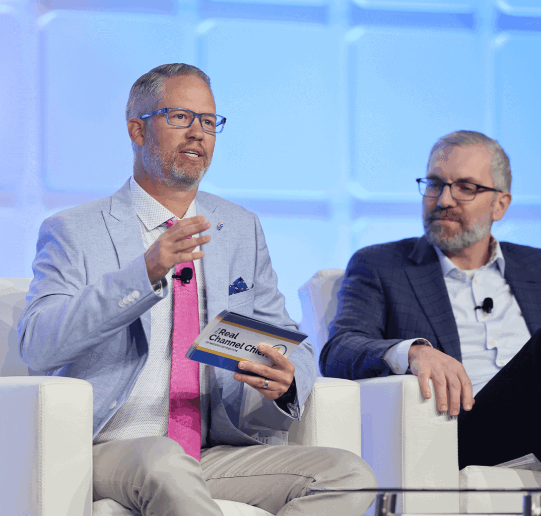 Two men in suits sit on a white stage panel. One man, holding a “Chief Channel Chief” card, speaks while gesturing with his hand. The other listens attentively. Both wear glasses; the background is blue and geometric.
