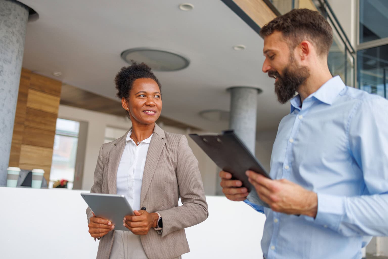Two business professionals, a woman in a beige suit holding a tablet and a man in a blue shirt holding a clipboard, smile and talk while standing in a modern office space with large windows and pillars.