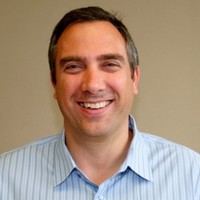 A man with short dark hair, wearing a light blue striped collared shirt, smiles in front of a plain beige background—reflecting confidence and empowerment, ideal for a 2025 Year in Review or Technology Advisor Enablement theme.