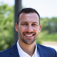 A man with short dark hair and a trimmed beard is smiling. He is wearing a blue suit jacket and white shirt. The outdoor background with greenery suggests a bright, sunny day—perfect for discussing Technology Empowerment in 2025 Year in Review.