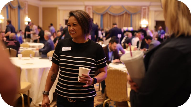 A woman with short hair and a striped black shirt smiles while holding a cup at a busy indoor event, with people seated and mingling in the background.