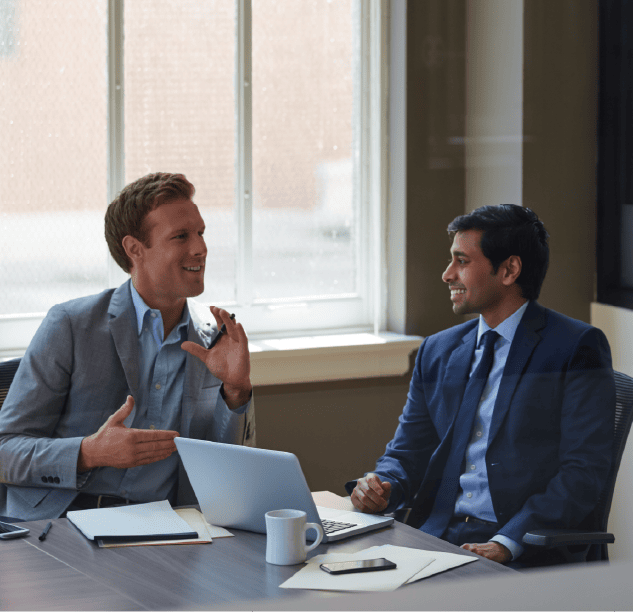Two men in business suits sit at a table in an office, smiling and talking. Laptops, notebooks, a coffee mug, and a smartphone are on the table—a scene reminiscent of collaborative learning at Telarus University. Sunlight streams in behind them.