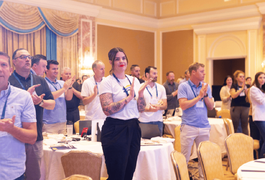A group of people stand and clap in a well-lit, elegant conference room with round tables and gold decor, appearing to give a standing ovation during a Telarus University Education Event or presentation.