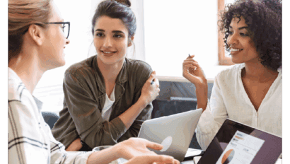 Three women sit at a table with laptops, smiling and talking. One gestures as they discuss effective AI conversions with results, suggesting a friendly, collaborative work meeting. A tablet displaying a chart is visible in the foreground.