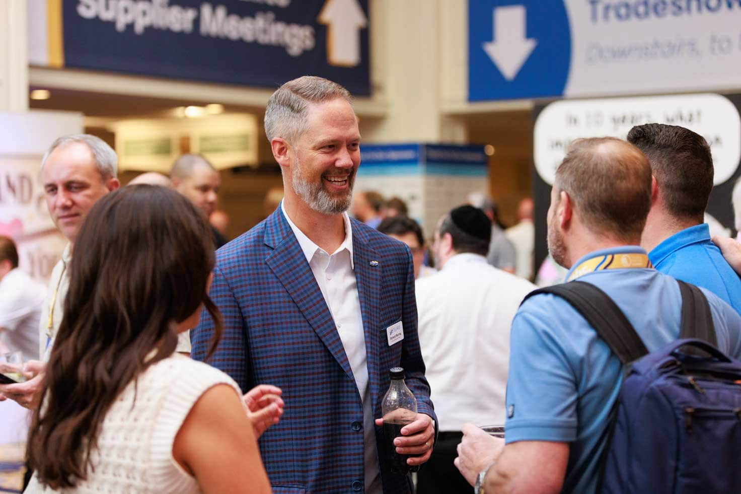 A man in a checked blazer smiles and converses with three people at a busy indoor event or tradeshow, as Telarus Tools signage and other attendees are visible in the background.