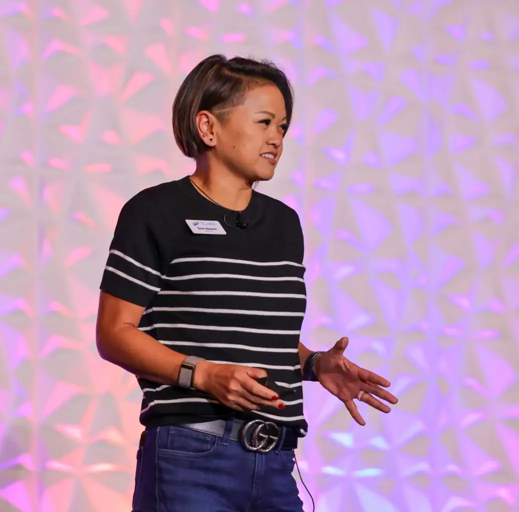 A person with short hair, wearing a striped black shirt and jeans, gestures while speaking on stage about Artificial Intelligence against a textured backdrop lit with pink and purple lights.