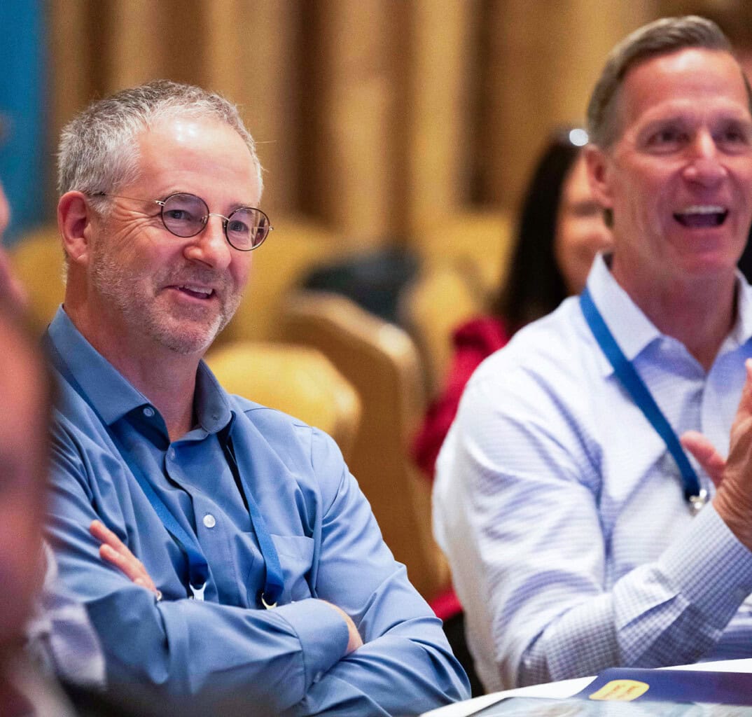 Two men wearing collared shirts and Telarus lanyards sit side by side at a table, smiling and clapping, during an indoor event. As technology services distribution allies, they celebrate success with others in the blurred background.