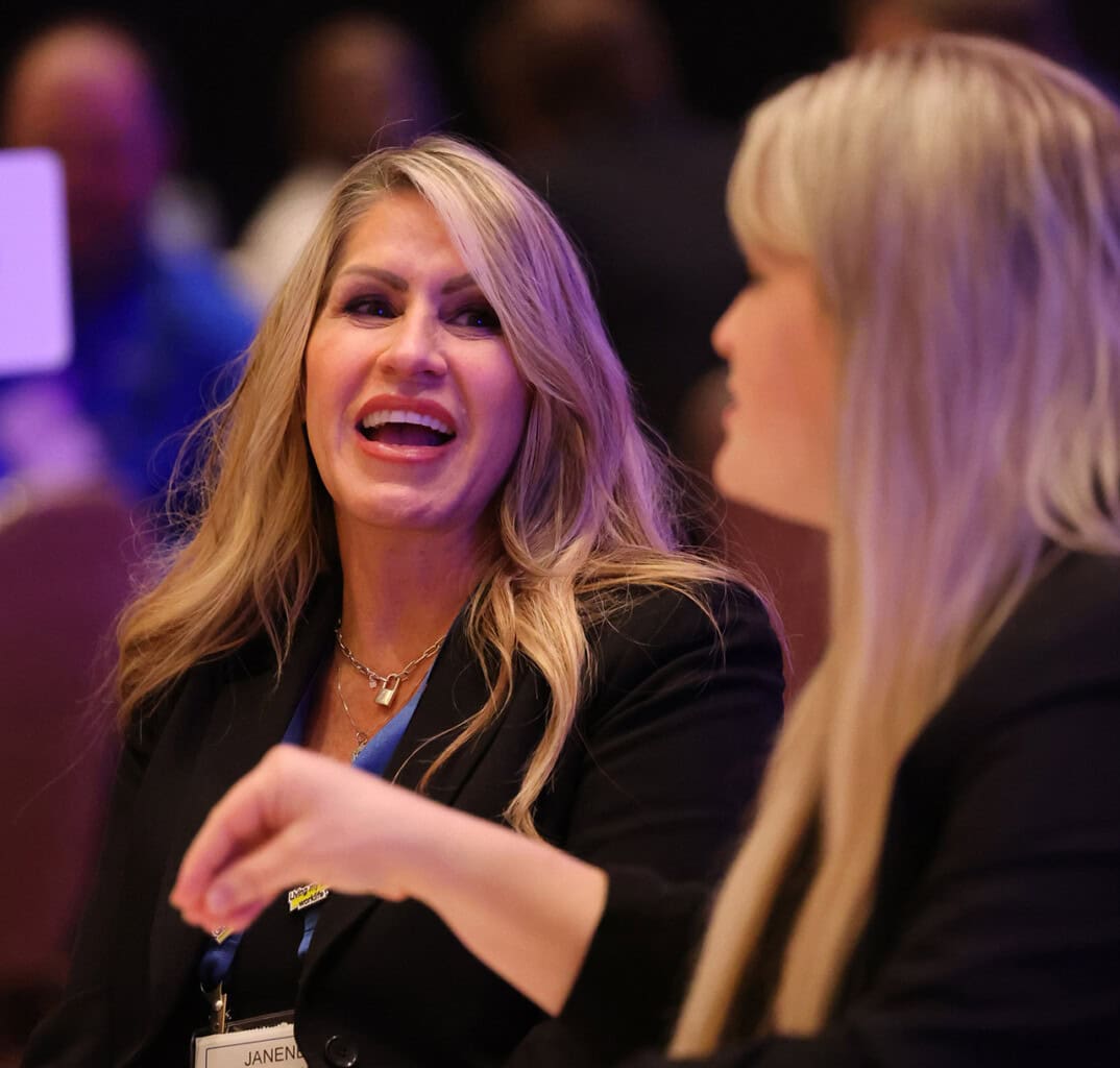 Two women with long blonde hair, dressed in black business attire, sit and talk animatedly at a conference. As Telarus technology services distribution allies, they engage in lively conversation, networking and sharing insights.