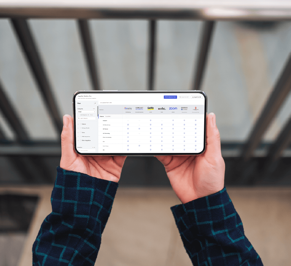Person holding a smartphone displaying a comparison chart of various online meeting platforms and Sales Applications, like Zoom and GoTo Meeting, with features listed and checkmarks highlighting their availability.