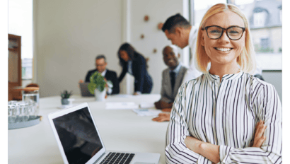 Smiling woman in glasses stands with arms crossed at a conference table with a laptop, while four colleagues discuss Microsoft AI strategy in the background in a bright, modern office.