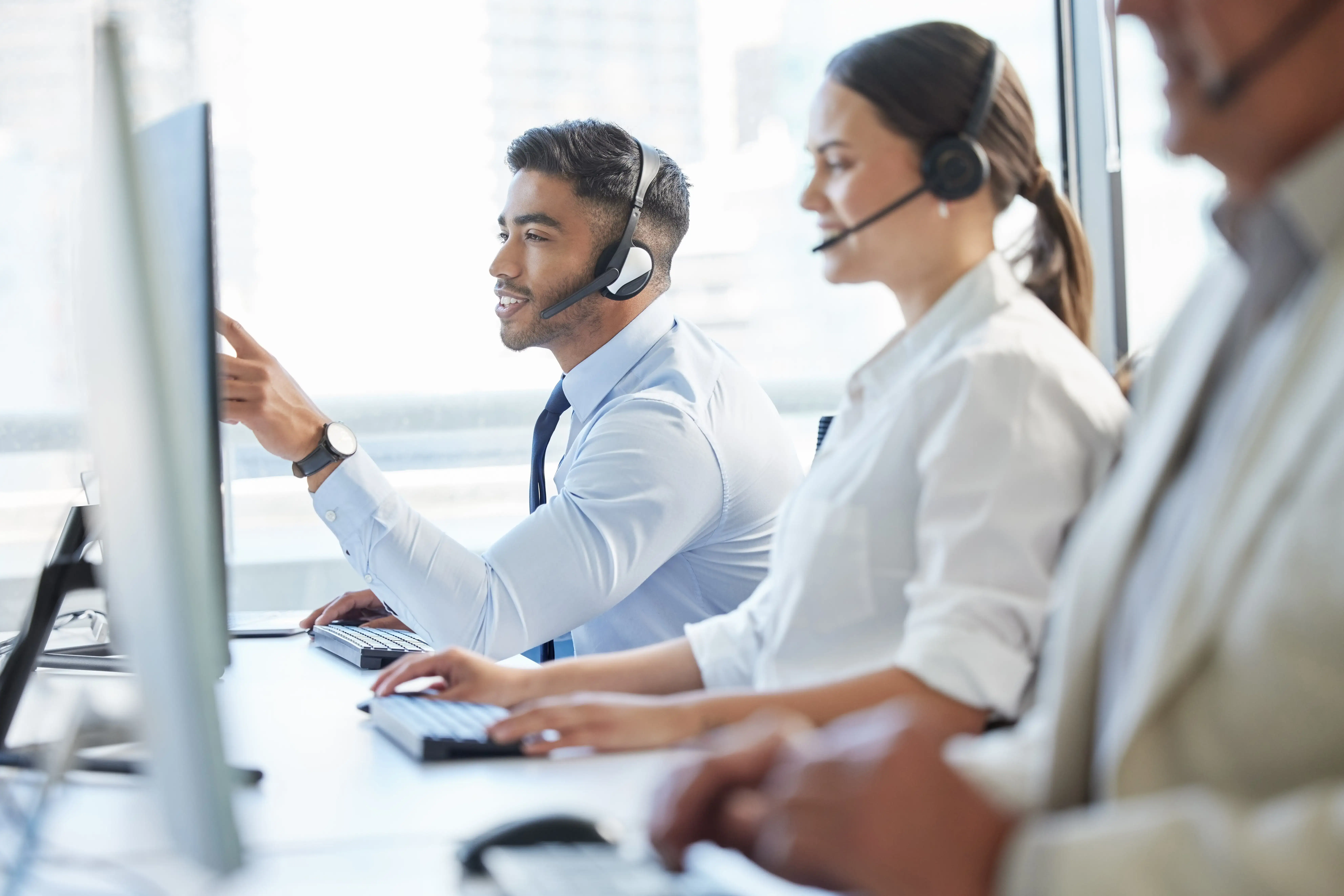 Three support call center employees wearing headsets work at their computers. The man in the foreground points at his monitor while speaking, and two colleagues beside him focus on their screens. Bright office environment.