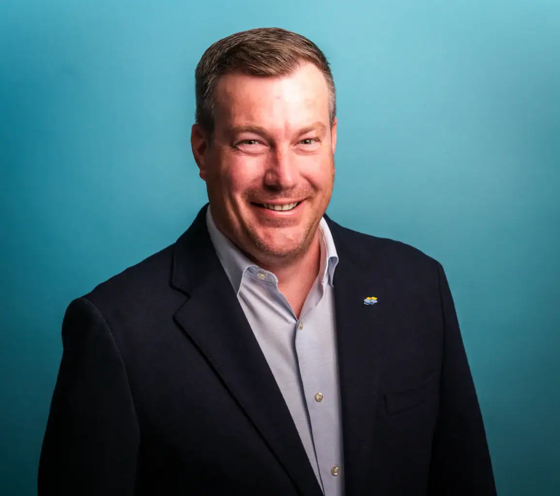 A man with short brown hair and a trimmed beard is smiling at the camera. He is wearing a navy blazer over a light blue shirt, with a blue pin on his lapel—subtly suggesting the hidden cost of style. The background is a solid light blue.