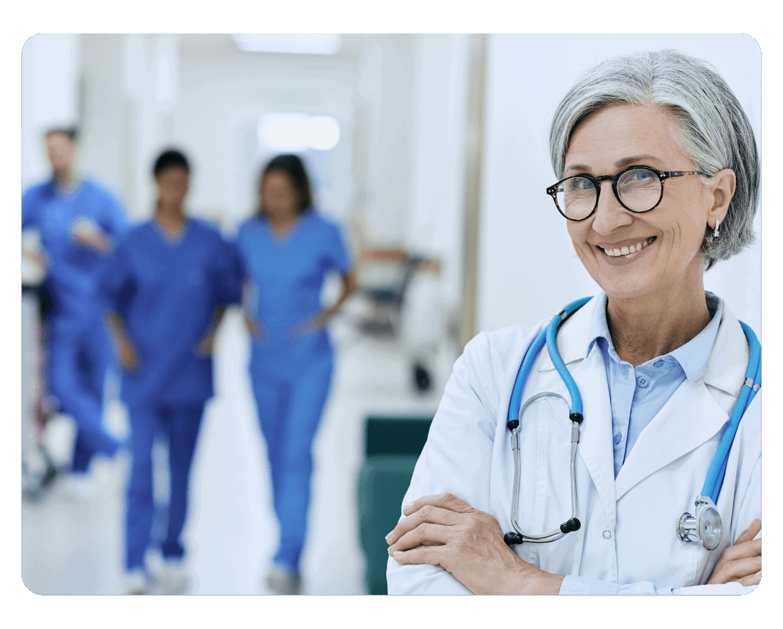A smiling older female doctor with gray hair and glasses, wearing a white coat and stethoscope, stands in the foreground. In the blurred hospital hallway behind her, three medical professionals discuss recent regulatory updates in blue scrubs.