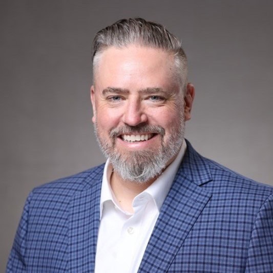Smiling man with short gray hair and beard, wearing a blue plaid suit jacket and white shirt, standing in front of a plain gray background—ready to share insights on regulatory updates and compliance trends.