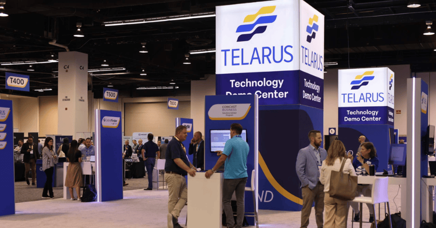 People walk and interact at a technology expo with large "Telarus Technology Demo Center" signs, booths, and digital displays visible throughout the convention hall.