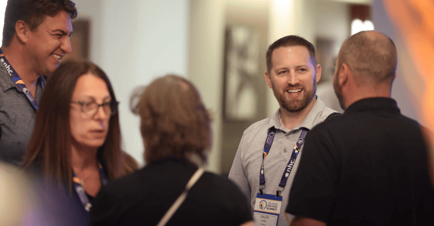 Five people stand in a group, talking and smiling at an indoor event. One man in the center wears a badge and blue lanyard. The background is softly blurred.