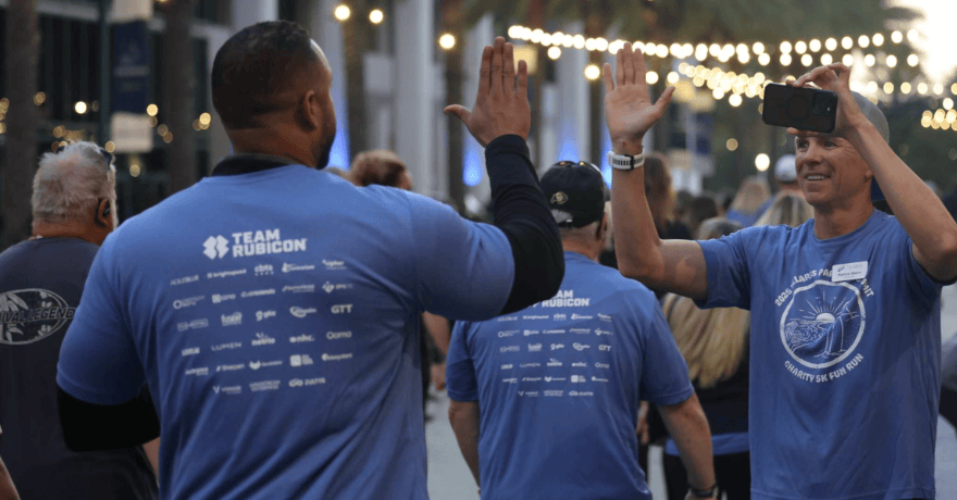 Two people in blue "Team Rubicon" shirts high-five at an outdoor event, with one person holding a phone. String lights and other participants are visible in the background.