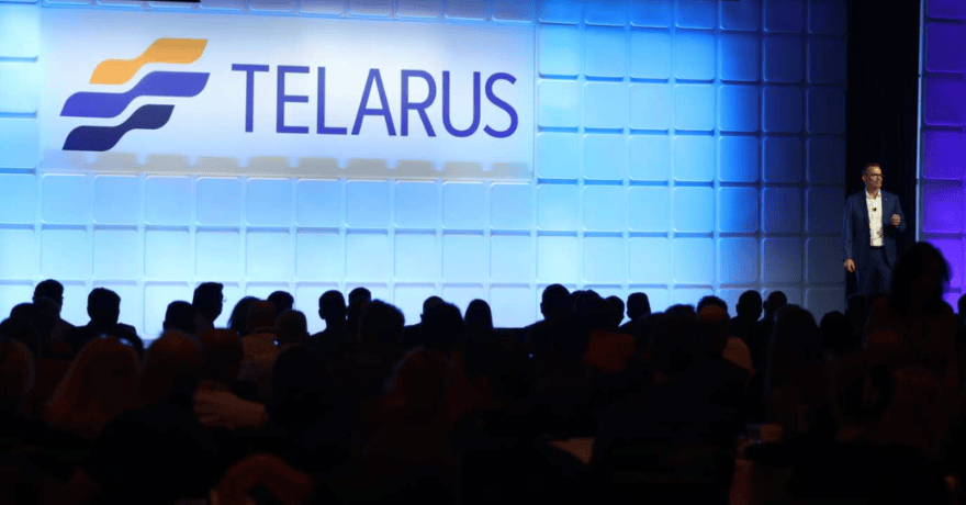A person stands onstage near a large sign reading "TELARUS" as an audience watches. The stage backdrop features a blue-tinted grid pattern.
