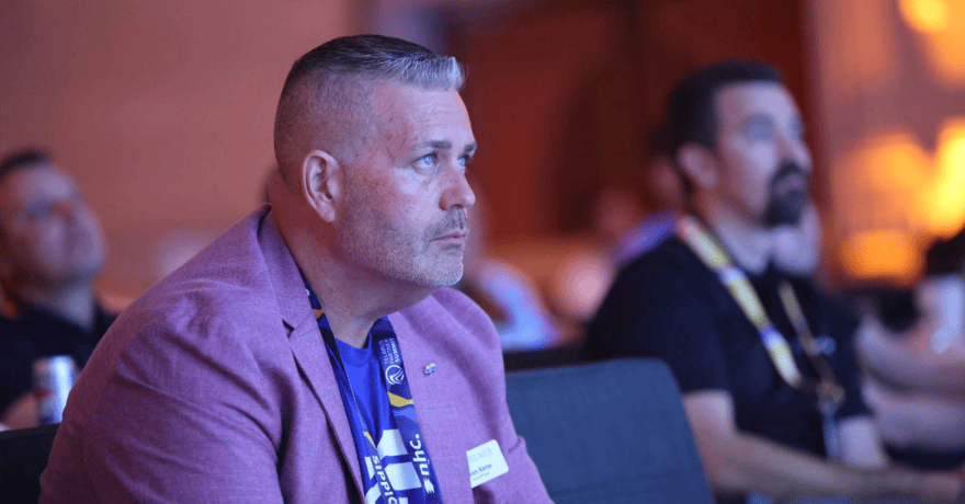 A man in a pink blazer sits attentively in an audience, looking forward. Other people are blurred in the background, suggesting a conference or event setting. Blue and warm lighting fill the scene.