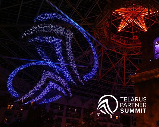 Hundreds of blue and purple drones form the Telarus logo in the air at night inside a large glass atrium, with a red-lit structure in the background and the Telarus Partner Summit 2026 logo at the bottom right—capturing a spirit of multi-solution growth and reflecting rising AI demand.